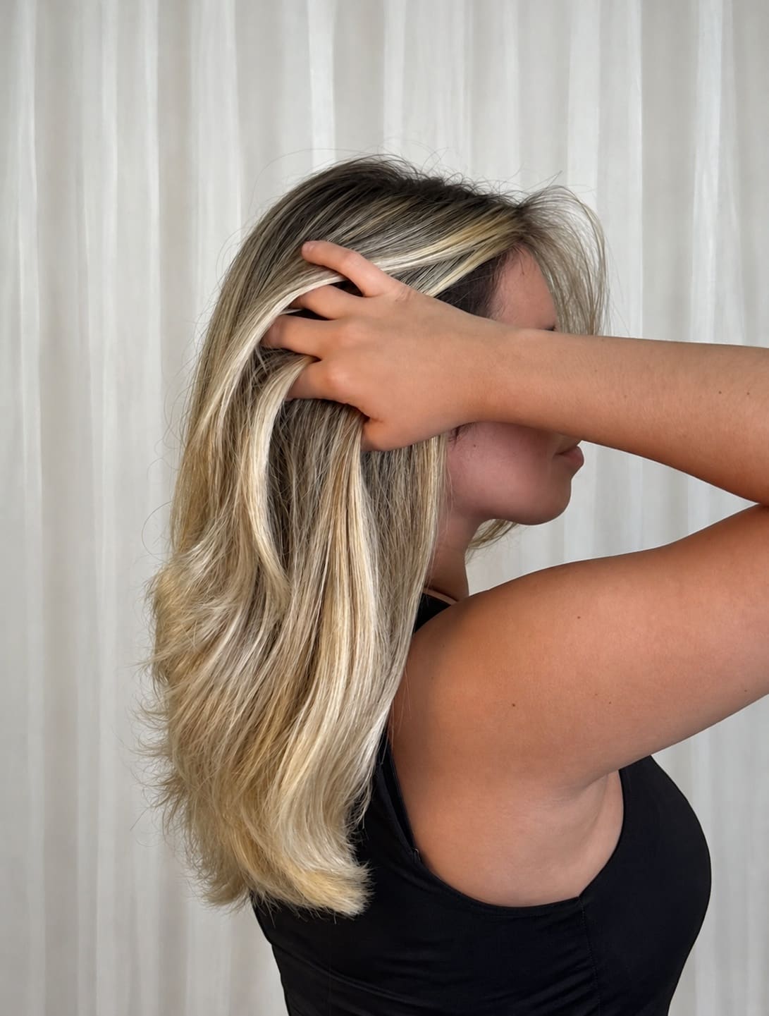 Woman with long blonde highlighted hair in side profile against white curtain backdrop