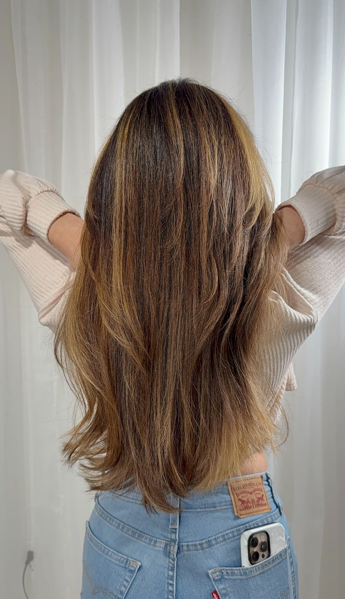 Woman with long layered brunette hair with caramel highlights, back view, wearing cream sweater and jeans