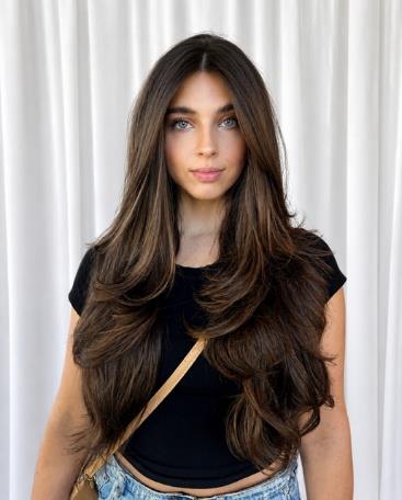 Woman with long dark brown wavy hair wearing black top against white curtain background