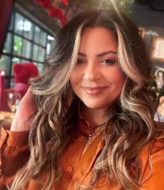 Woman with long layered brunette hair with blonde highlights wearing an orange button-up shirt