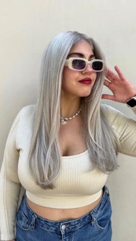 Woman wearing white sunglasses, gray long hair, pearl necklace, cream crop top, and denim jeans