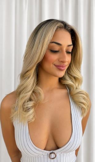 Woman with long blonde wavy hair wearing white ribbed sleeveless top against white curtain background