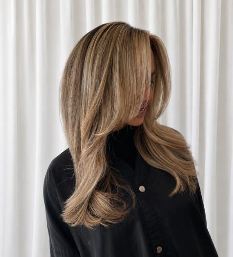Woman with long, layered blonde hair wearing a black button-up shirt against a white curtain backdrop